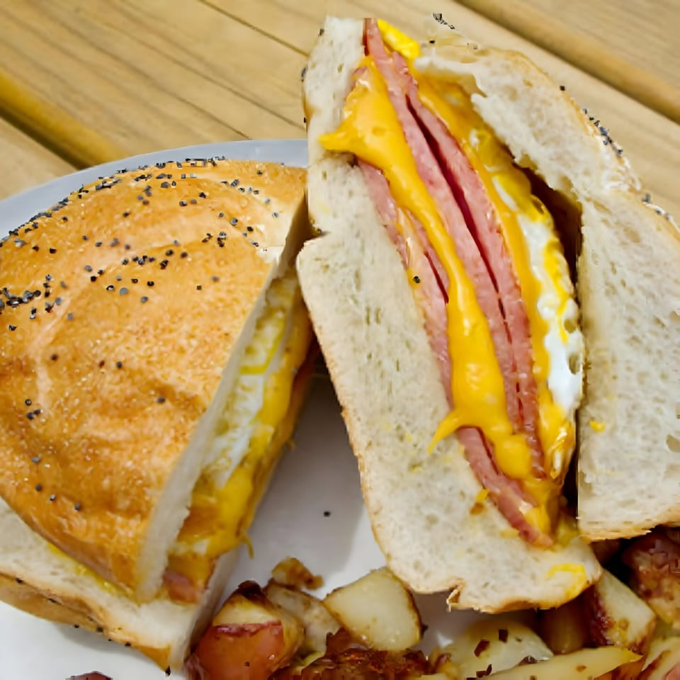A sliced deli sandwich with ham, melted cheese, and egg, served with home fries.