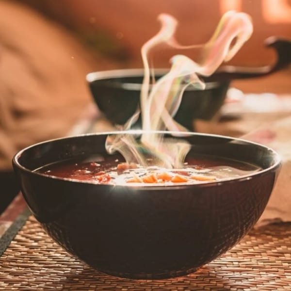 Steaming hot soup with vegetables in a black bowl on a woven placemat at Agamie Gourmet.