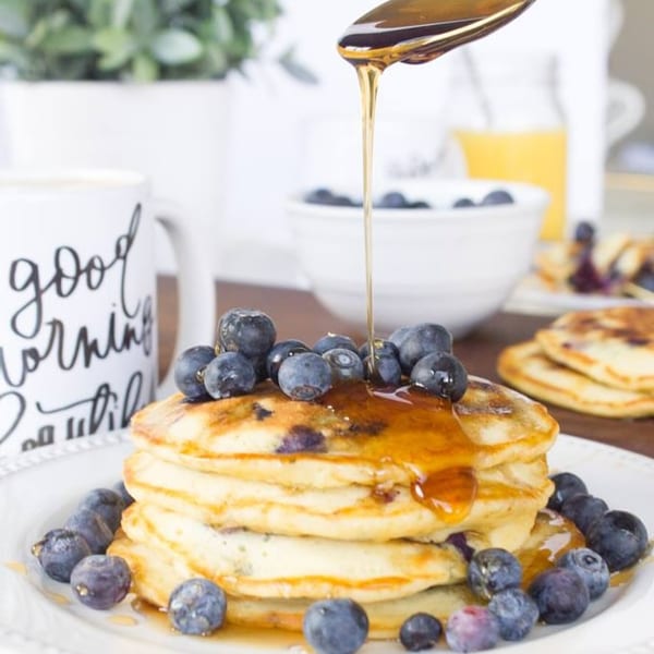 Syrup being poured over a stack of blueberry pancakes, with a "good morning" mug nearby.